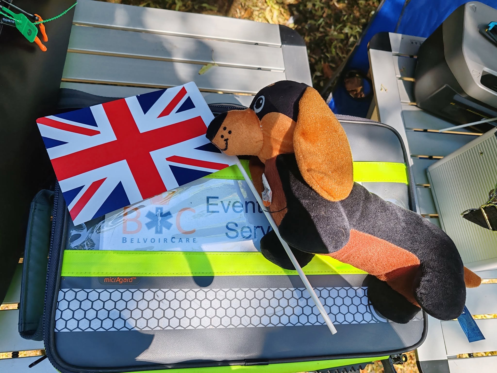 Mr Shrimpy, BelvoirCare's friendly mascot — a plush dachshund holding a Union Jack flag, resting on a branded BelvoirCare medical kit bag.