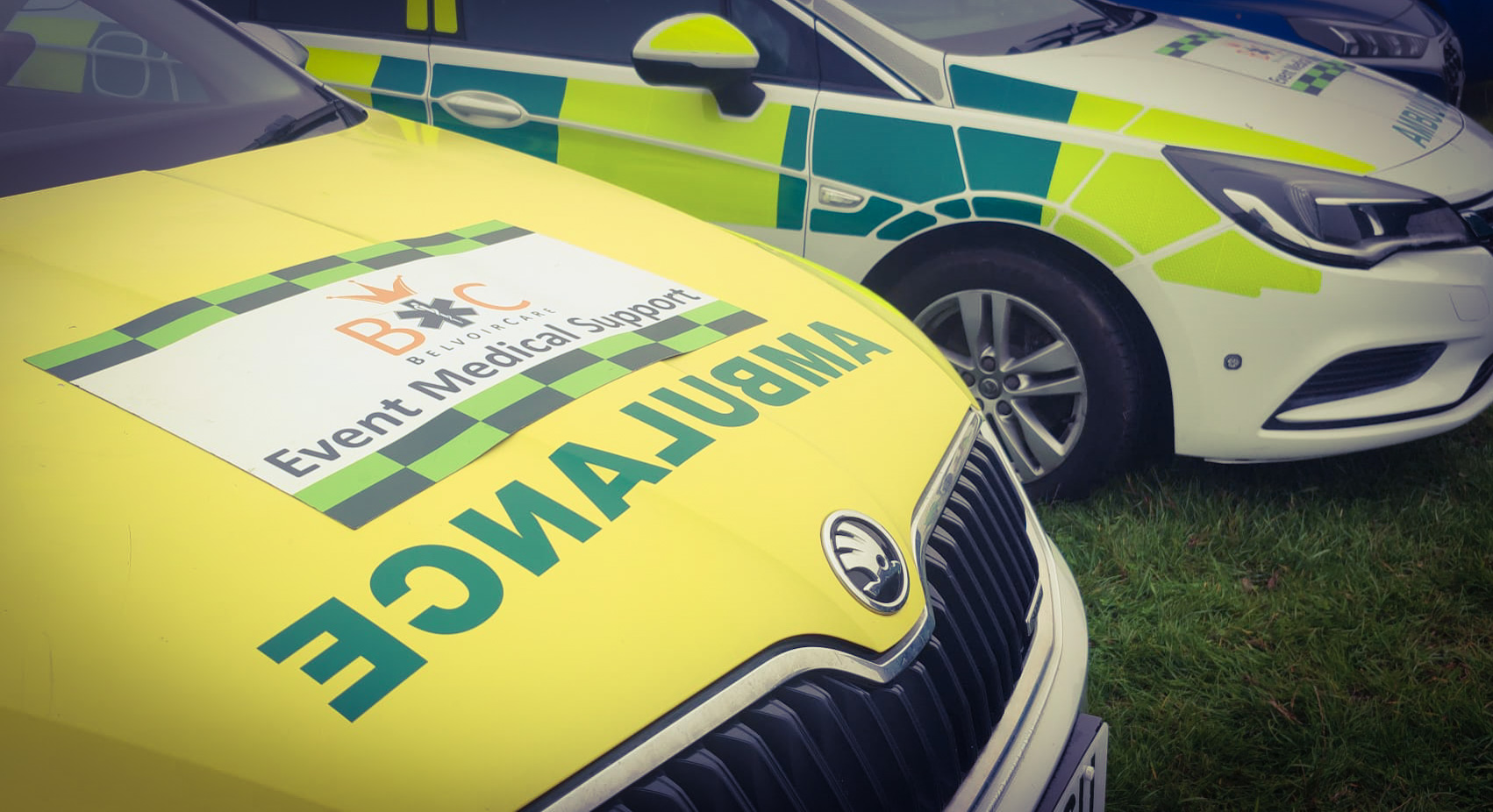 A BelvoirCare event ambulance branded 'Event Medical Support' parked alongside a green-and-yellow rapid-response car.