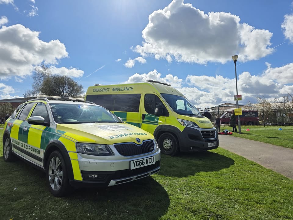 A BelvoirCare emergency ambulance and ambulance-responder car on a sunny day at an outdoor event.